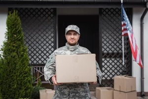 front view of military serviceman with cardboard box standing near house with american flag